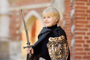 Garçon blond en cape noire avec épée et bouclier au lion devant un mur de briques, prêt pour son anniversaire chevalier