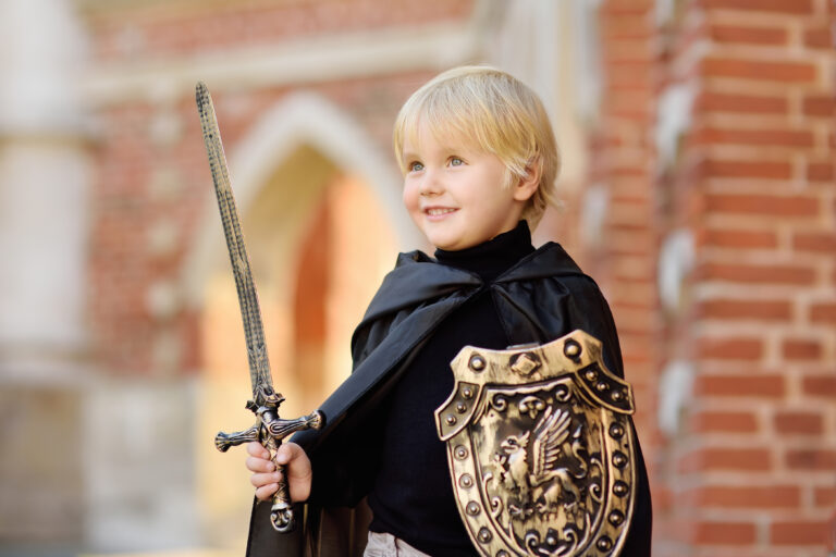 Garçon blond en cape noire avec épée et bouclier au lion devant un mur de briques, prêt pour son anniversaire chevalier