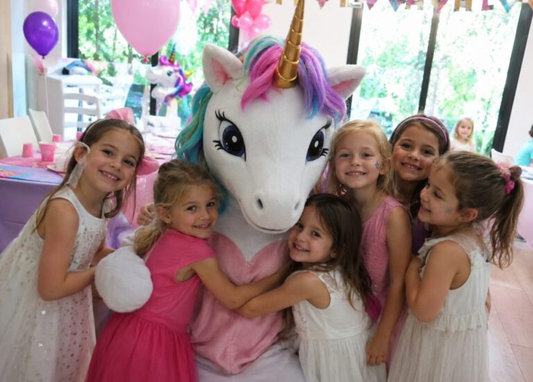 Anniversaire licorne : sept fillettes en robes pastel serrées autour d'une grande peluche licorne arc-en-ciel