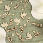 Confettis de table en bois clair : colombes, branches d'olivier et mot Baptême dispersés sur un voile sauge