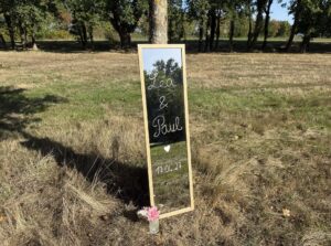 Miroir d'accueil de mariage à cadre bois posé dans l'herbe, prénoms Léa et Paul et date écrits en blanc