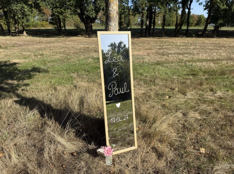 Miroir d'accueil de mariage à cadre bois posé dans l'herbe, prénoms Léa et Paul et date écrits en blanc