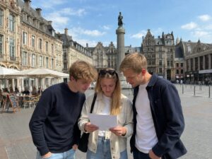 Trois jeunes adultes lisent la feuille d'un jeu de piste sur la Grand-Place de Lille, façades flamandes