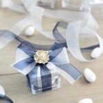 Cube transparent noué d'un ruban en organza bleu marine et blanc avec fleur crème, entouré de dragées blanches