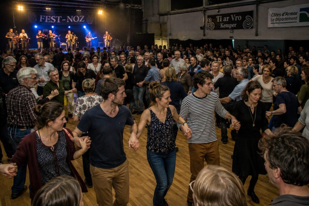 Danseurs se tenant par la main en ronde devant des musiciens sur scène lors d'un fest-noz breton en salle