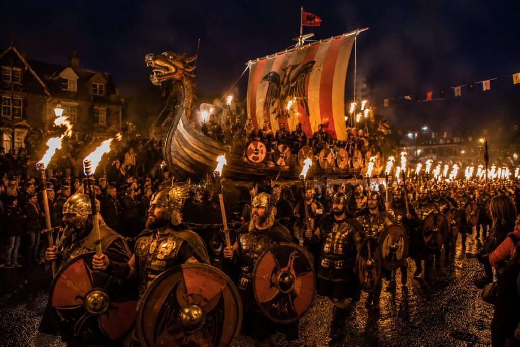 Guerriers vikings casqués portant boucliers et torches devant une galère à proue de dragon lors d'Up Helly Aa
